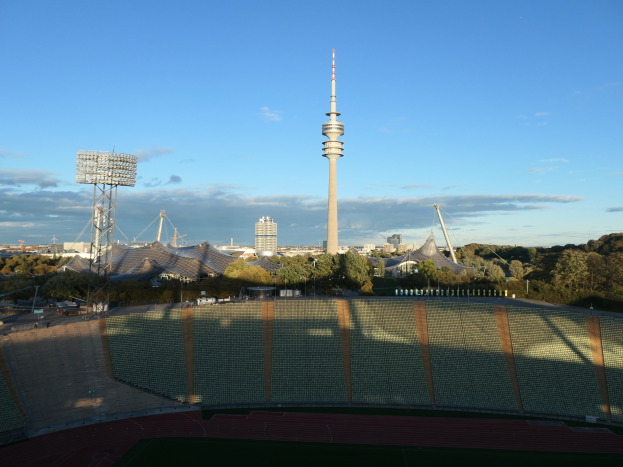 Das Olympische Stadion in Berlin, Deutschland, mit dem Fernsehturm im Hintergrund, umgeben von Bäumen, Gebäuden und Lichtern unter einem bewölkten Himmel.