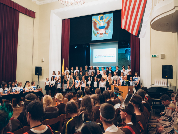 Gruppe von Menschen vor sitzendem Publikum bei einem Event mit Podium, Mikrofon, Flaggen, Lautsprechern, einem Bildschirm mit Text und Bild sowie Vorhängen und einem Kronleuchter.