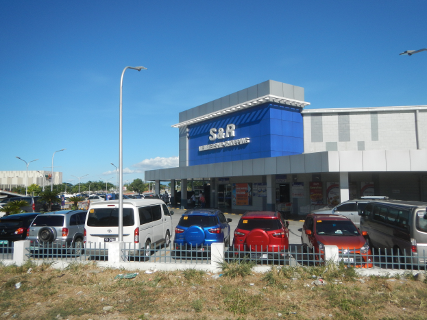 Ein Autohaus mit parkenden Fahrzeugen vorne, umgeben von einem Metallzaun, Gras, Pflanzen, Bäumen, Straßenlaternen.