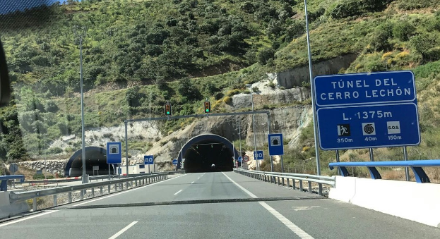 Auto fährt durch einen Tunnel auf einer Straße mit Geländern, Schildern, Pfählen, Verkehrszeichen, Bäumen, Felsen und einem klaren blauen Himmel.