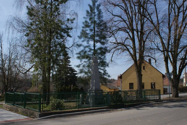 Denkmal für Holocaust-Opfer umgeben von einem Metallzaun, Pflanzen, Gras, Bäumen, Häusern, einem Straßenpfahl und einem bewölkten Himmel.