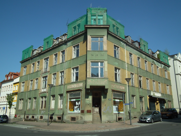 Altes Gebäude mit grüner Fassade an einer Straßenecke, umgeben von Straßenmasten, Straßenlaternen, Kraftfahrzeugen, Bäumen und einem klaren blauen Himmel.