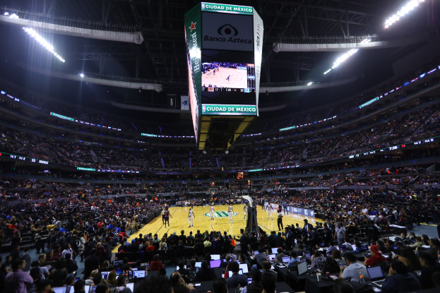 Basketballspiel in einer großen Arena mit Zuschauern, die auf Stühlen sitzen und stehen, in der Nähe von Tischen mit Laptops und Flaschen, Basketballkorb und Anzeigebildschirm im Hintergrund.