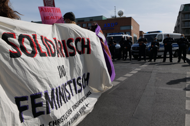 Eine Gruppe von Menschen marschiert mit einem Banner mit der Aufschrift 'Solidarität und Feminismus' die Straße entlang, mit geparkten Fahrzeugen am Straßenrand und Gebäuden im Hintergrund unter einem klaren blauen Himmel.