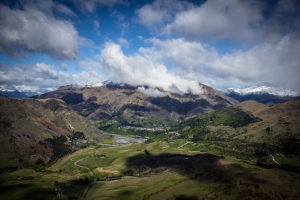 Ein atemberaubender Blick von der Spitze eines Berges in Queenstown, Neuseeland, der saftiges Grün, Bäume, eine gewundene Straße und einen Himmel voller weißer, flauschiger Wolken zeigt.