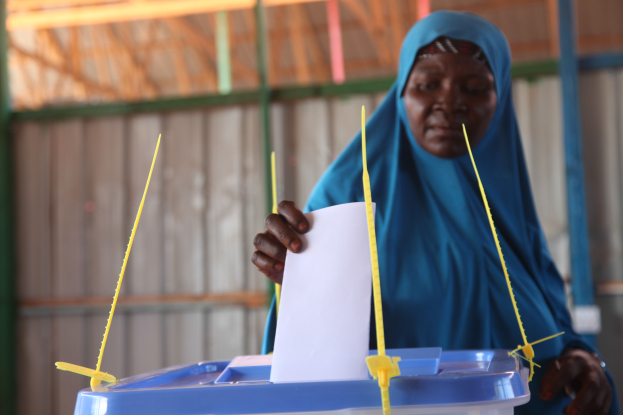 Eine Frau mit einem blauen Kopftuch steht und hält ein Blatt Papier in der Hand, vor ihr steht eine Kiste mit Stöcken, im Hintergrund eine Wand und ein Schuppen.