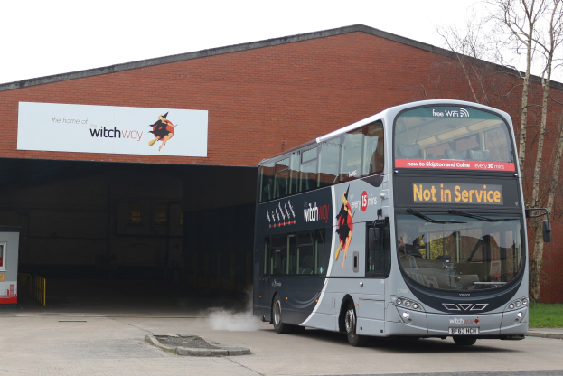 Doppeldeckerbus mit Rauchwolke an einem Gebäude vorbeifahrend, mit Gras, Bäumen und einem blauen Himmel im Hintergrund, sowie einem sichtbaren Fahrzeugkennzeichen am Bus.