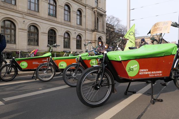Gruppe von Fahrrädern, die entlang einer Straße geparkt sind, mit einer Person in der Nähe, vor einem Gebäude, Bäumen und einem klaren Himmel, mit einem Banner für eine Fahrrad-Sharing-Kampagne.