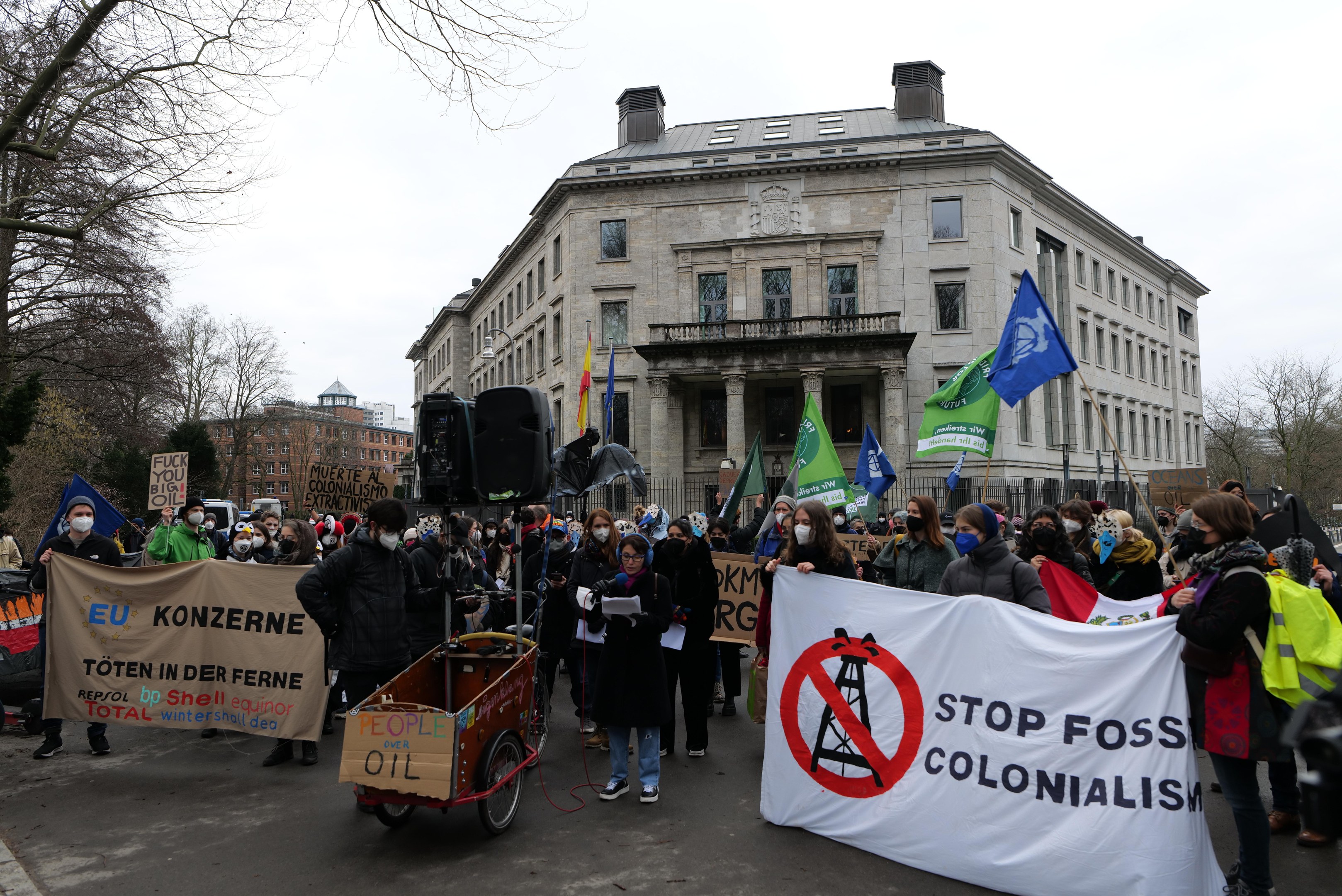 Eine große Gruppe von Menschen marschiert mit Schildern und Fahnen eine Straße entlang, mit einem Fahrzeug im Vordergrund, und protestiert gegen fossile Brennstoffe; Bäume, Gebäude mit Fenstern und ein klarer blauer Himmel sind im Hintergrund zu sehen.
