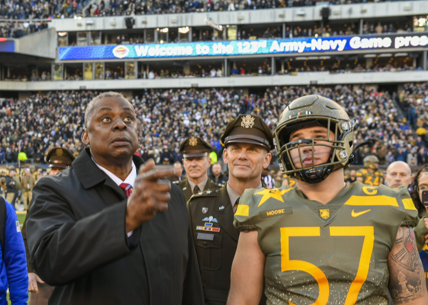 Ein Mann im Anzug und Krawatte steht neben einem Football-Spieler in Uniform und Helm, umgeben von Menschen in Team-Uniformen und Kappen, mit einer Stadionmenge und einer Tafel mit der Aufschrift "Oregon Football Coach Mike McCarthy und Oregon QB Marcus Mariota" im Hintergrund.