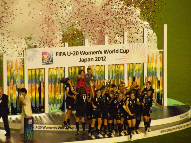 Gruppe von Frauenfußballspielerinnen in schwarzen Uniformen, die auf einer Bühne feiern und von einer Menge umgeben sind, mit einer Tafel und dekorativen Elementen im Hintergrund.