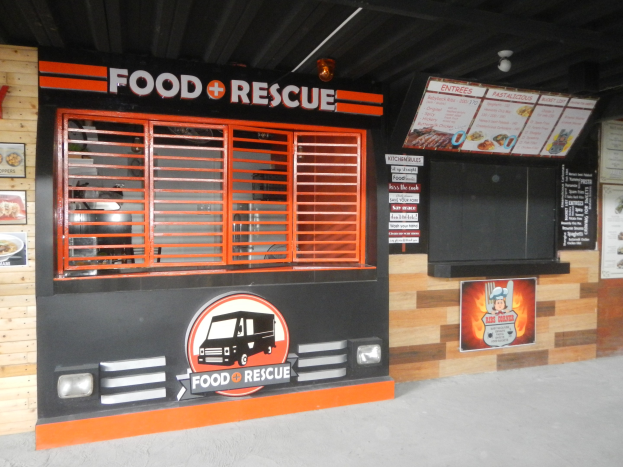 Ein Foodtruck vor einem Gebäude mit einer "Food Rescue"-Tafel geparkt, mit einem Fenster, einer Tür, an der Wand montierten Tafeln mit Text und Bildern und Deckenleuchten.