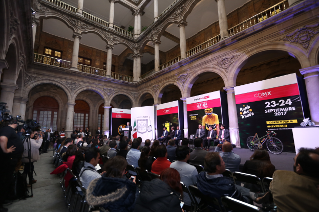 Eine Gruppe von Menschen, die an einem Tisch mit einem Podium und einem Mikrofon vor einer Menge, Kameras und Transparenten sitzt, während einer Pressekonferenz für das CDMX 2017-Event.
