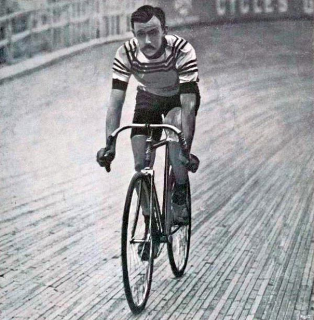 Schwarz-weiß-Foto eines Mannes, der auf einer Bahn ein Fahrrad fährt, mit einem Zaun und einer Tafel im Hintergrund.