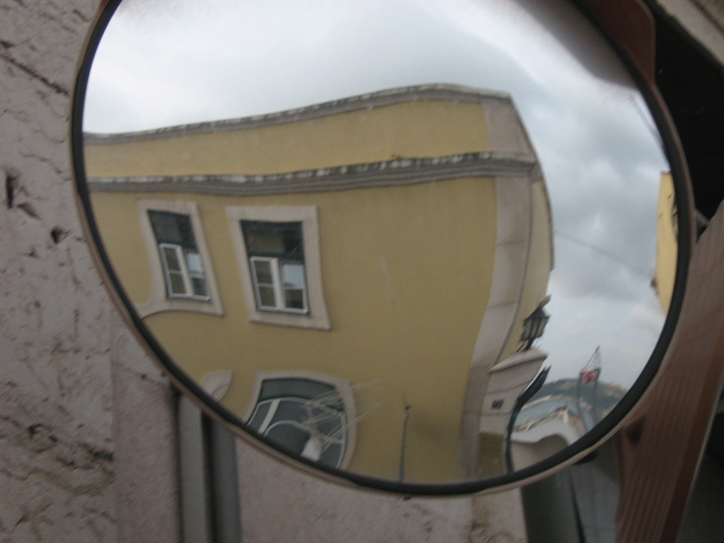 Ein Straßenspiegel, der Gebäude, Lichter und den Himmel reflektiert, mit einer Wand im Hintergrund.