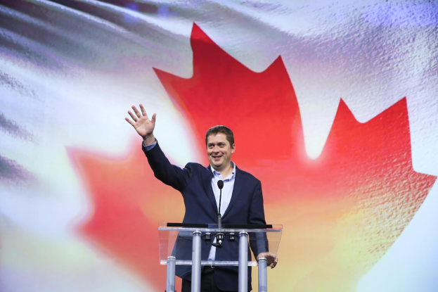 Der kanadische Premierminister Alex Salmond steht hinter einem Rednerpult mit Mikrofonen, lächelt vor einer kanadischen Flagge und hat einen Bildschirm im Hintergrund.