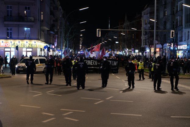 Eine Gruppe von Menschen, die über eine nächtliche Stadtstraße gehen, mit Straßenlaternen, Gebäuden, Bäumen, Verkehrsampeln, Fahrzeugen und einem Banner im Hintergrund.