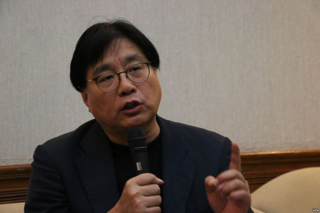 Ein Mann im Anzug und mit Brille spricht in ein Mikrofon, während er eine Rede hält, mit einer Wand im Hintergrund.