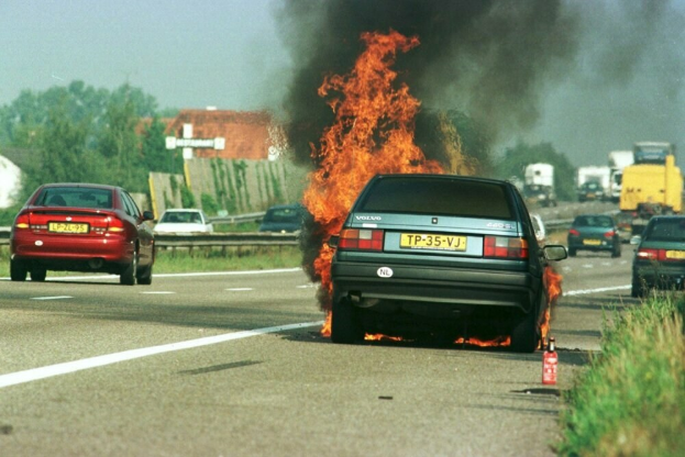 Ein Auto in Flammen auf dem Seitenstreifen, umgeben von anderen Fahrzeugen, mit Bäumen, Gebäuden und einem klaren blauen Himmel im Hintergrund und Gras mit einem Feuerlöscher auf der rechten Seite.