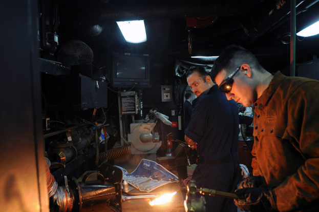 Zwei Männer mit Brille arbeiten an einem Metallteil in einer Werkstatt, einer verwendet ein Schweißgerät, mit verschiedenen Werkzeugen und Geräten auf dem Tisch und einem Fernseher und Beleuchtung im Hintergrund.