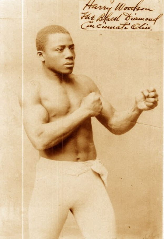 Altes Schwarz-Weiß-Foto eines Boxers im Ring mit Handschuhen und Shorts, der mit konzentriertem Blick und entschlossener Miene steht.