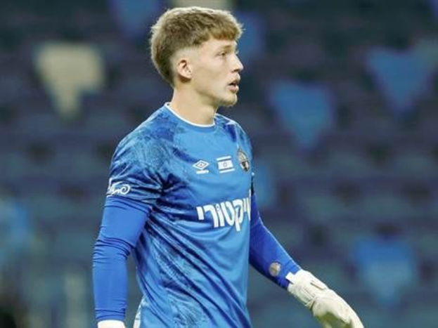 Ein junger Mann in einem blauen Torwarttrikot steht auf einem Fußballfeld in einem blauen T-Shirt und weißen Handschuhen während eines Spiels.