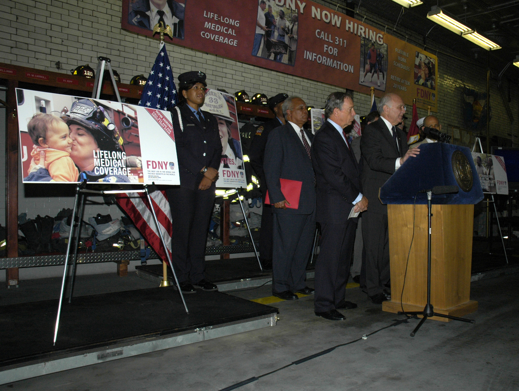 Eine Gruppe von Menschen in der Nähe eines Podiums mit Mikrofonen, Schildern auf Ständern, Fahnen und Schildern an der Wand, alles unter hellen Lichtern.