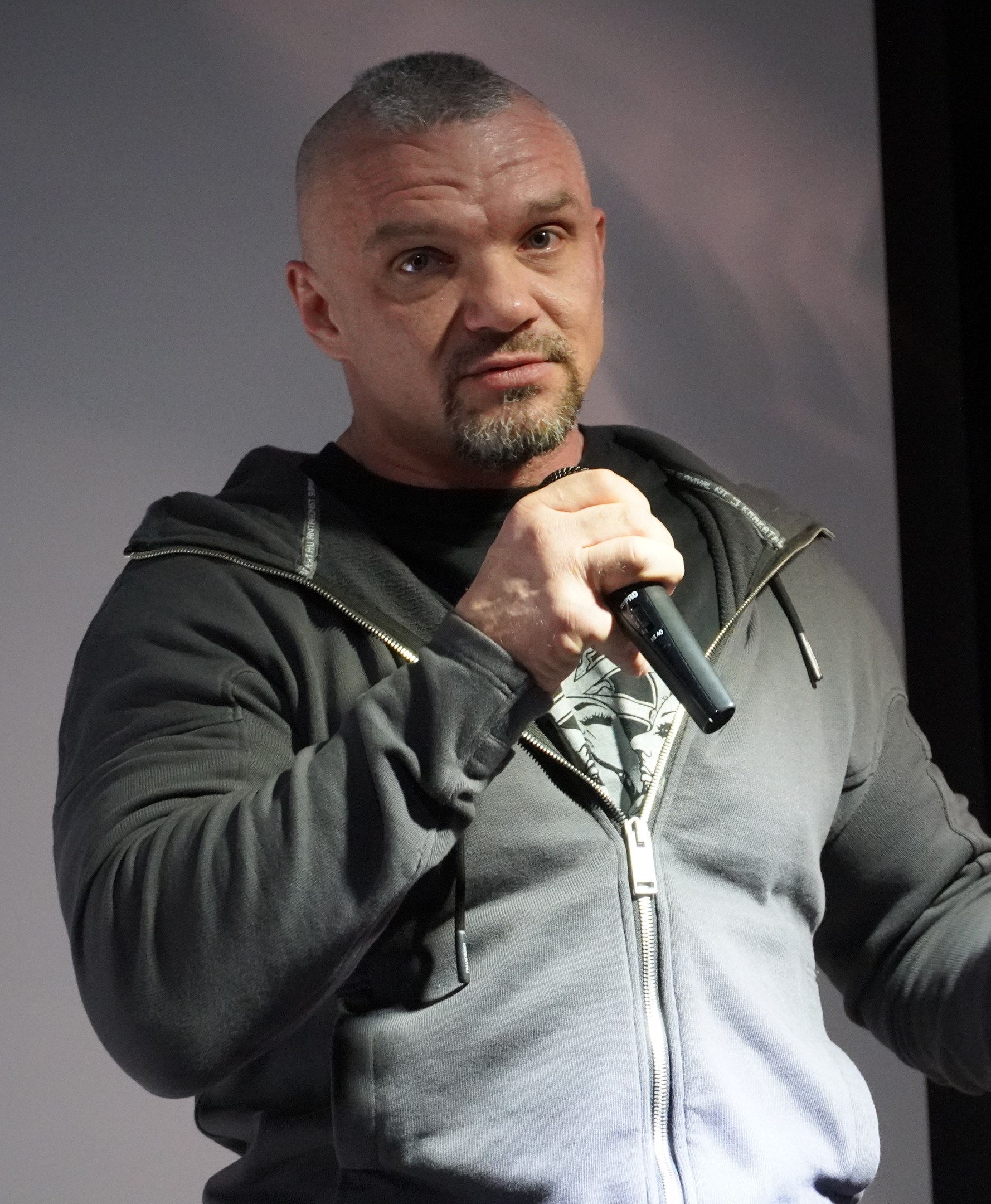 Ein Mann in einer schwarzen Kapuzenjacke und einem schwarzen Anzug mit einem weissen Hemd, der ein Mikrofon halt, steht selbstbewusst vor einer Wand und scheint ein Komiker mit zurückgek├Ąmmtem Haar zu sein.