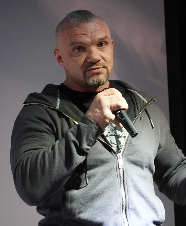 Ein Mann in einer schwarzen Kapuzenjacke und einem schwarzen Anzug mit einem weissen Hemd, der ein Mikrofon halt, steht selbstbewusst vor einer Wand und scheint ein Komiker mit zurückgek├Ąmmtem Haar zu sein.