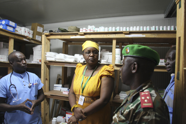 Vier Menschen in einer Apotheke: Eine Frau in einem gelben Kleid und grüner Mütze steht zwischen zwei Männern in blauen Hemden mit Stethoskopen, umgeben von Medikamentenflaschen und -schachteln auf der Theke und Regalen.