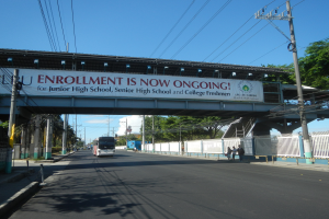 Ein Bus fährt unter einer Brücke mit einem Schild "Jetzt Anmelden" vorbei, während Menschen auf einem Fußweg darunter gehen, mit Strommasten und Drähten entlang der Straße und Bäumen und einem klaren blauen Himmel im Hintergrund.