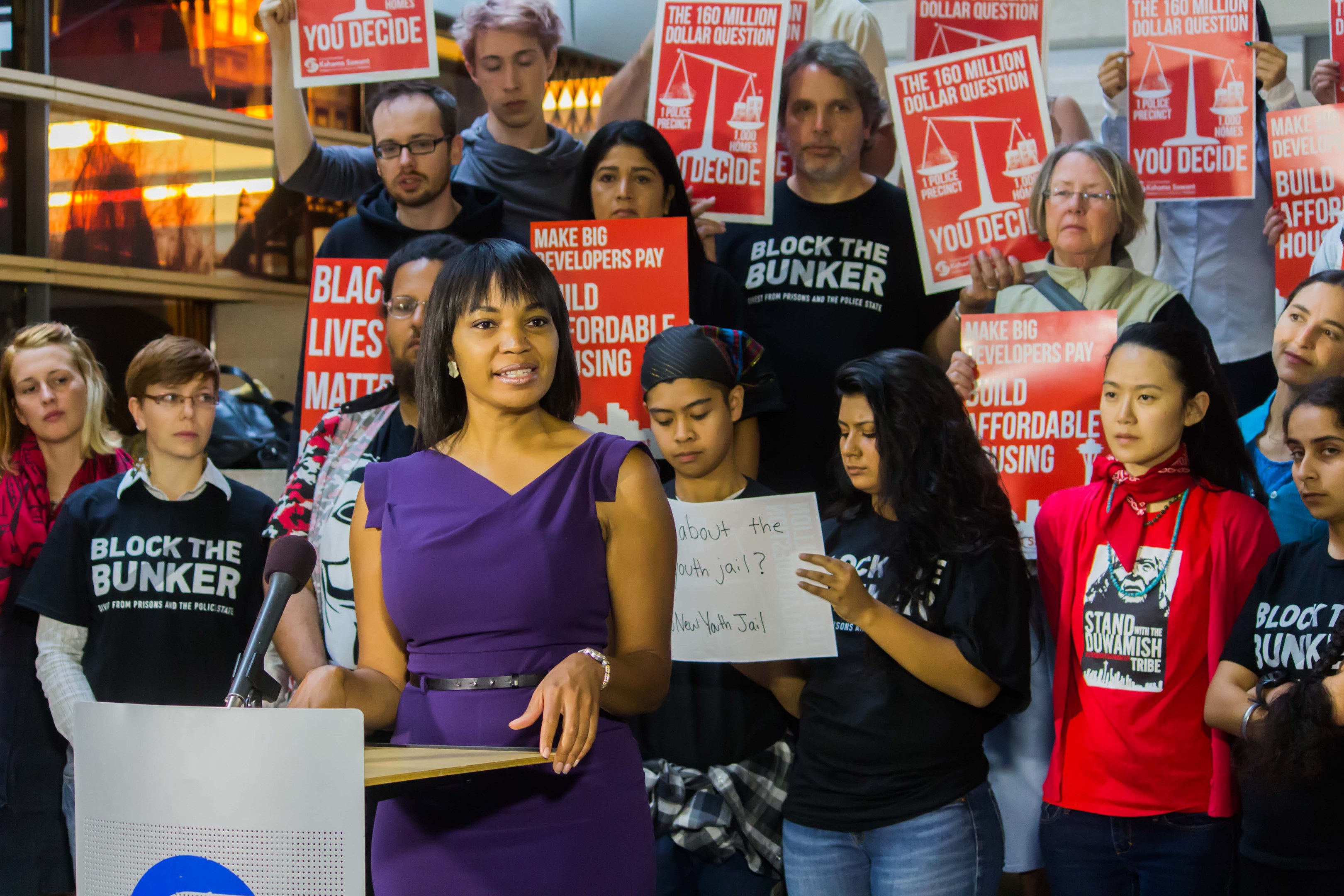 Eine Black-Lives-Matter-Aktivistin spricht an einem Rednerpult mit Mikrofon zu einer Menge mit Schildern, mit Scheinwerfern und Equipment im Hintergrund.