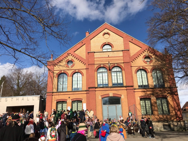 Eine große Gruppe von Menschen steht vor einem mehrstöckigen Gebäude mit Fenstern und Türen, umgeben von Bäumen unter einem bewölkten Himmel, wobei einige Teilnehmer Mützen tragen und Taschen tragen.