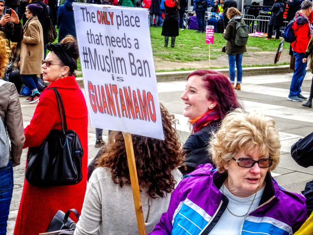 Eine Gruppe von Menschen steht auf dem Boden, einige halten Taschen, eine Frau hält ein Schild mit der Aufschrift "Der einzige Ort, der ein muslimisches Verbot benötigt, ist Guantanamo", vor einem Metallzaun, Gras, einem Schild, Pfosten, einem Gebäude mit Fenstern und Bäumen.