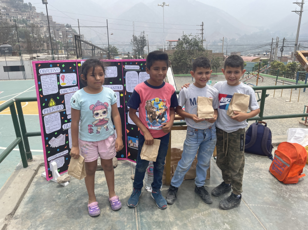 Gruppe von Kindern auf einem Basketballfeld mit Papiertüten in den Händen, mit Gegenständen auf dem Boden, einer Tafel mit Text links daneben und einem Zaun, Pfosten und Gebäuden im Hintergrund.