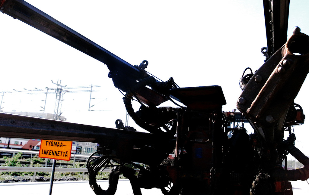 Eine Maschine im Vordergrund mit Strommasten, Kabeln, Barrieren, Pflanzen und Himmel im Hintergrund.