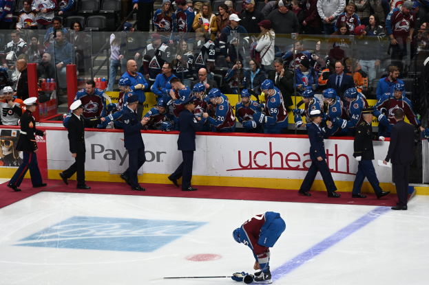 Hockey player on ice surrounded by teammates and opponents during a game, with spectators in the background and arena boards displaying the matchup "Colorado Avalanche vs San Jose Sharks - 3/24/15 NHL Pick, Odds, and Prediction".