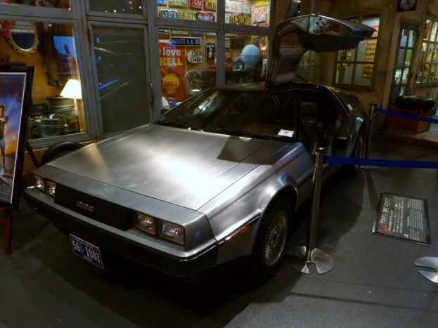 Ein DeLorean DMC-12 aus der Zurück in die Zukunft-Reihe in einem Museum ausgestellt, dessen Türen offen sind, umgeben von Informationsanzeigen, Bändern und verschiedenen Gegenständen.