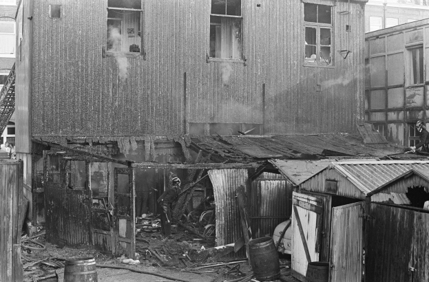 Ein Schwarz-Weiß-Foto eines durch Feuer beschädigten Gebäudes mit verstreuter Trümmern, darunter Fässer und hölzerne Gegenstände, sowie einige Menschen in der Nähe, mit anderen Gebäuden, einer Leiter und einem Pfahl im Hintergrund.