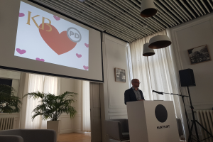 Ein Mann steht an einem Podium mit einem Mikrofon, gibt eine Präsentation vor einer Projektionswand, mit Stühlen, einer Pflanze, Fotorahmen, Vorhängen und Deckenleuchten im Hintergrund.