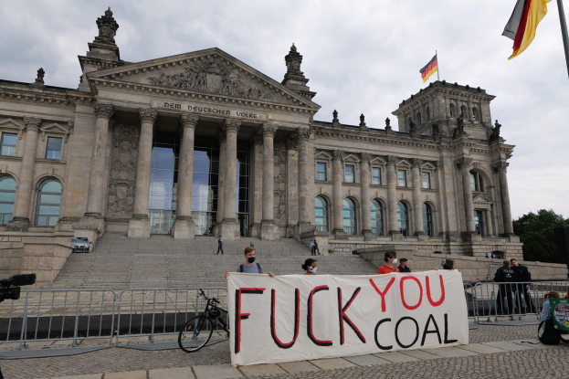 Eine Gruppe von Menschen hält ein Transparent mit der Aufschrift "Fuck You Coal" vor dem Reichstaggebäude in Berlin, Deutschland, mit Bäumen, einer Fahnenstange und einem bewölkten Himmel im Hintergrund.