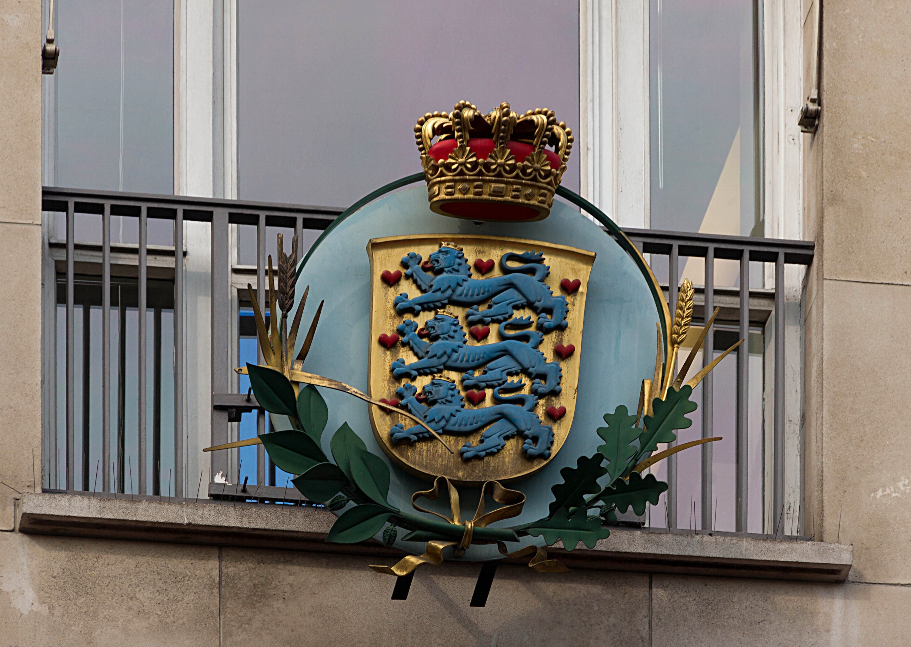 Wappen der britischen Königsfamilie an einer Gebäudefassade mit einer Krone und -blätter-Logo, Fenstern und Metallgittern im Hintergrund.