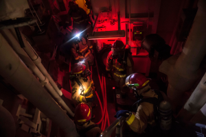 Eine Gruppe von Feuerwehrmännern in Helmen und Sauerstoffzylindern steht in einem dunklen Raum mit Rohren auf beiden Seiten, mit einer Maschine und anderen Gegenständen im Hintergrund, was auf einen Rettungseinsatz hinweist.