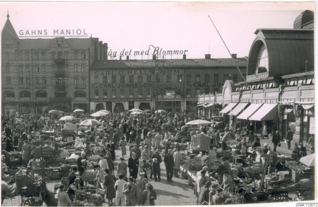 Ein belebter Markt im alten Stadtzentrum von Heidelberg mit Menschen an Tischen mit Blumentöpfen und Schirmen, umgeben von Gebäuden, Bäumen und einem klaren blauen Himmel.