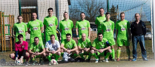 Eine Gruppe von Männern in grünen T-Shirts steht und hockt auf einem Fußballfeld mit einem Ball davor, ein Torpfosten rechts und ein Hintergrund aus Bäumen, Gebäuden, Polen, Fahrzeugen und Himmel, mit einer Zahlenfolge links unten.