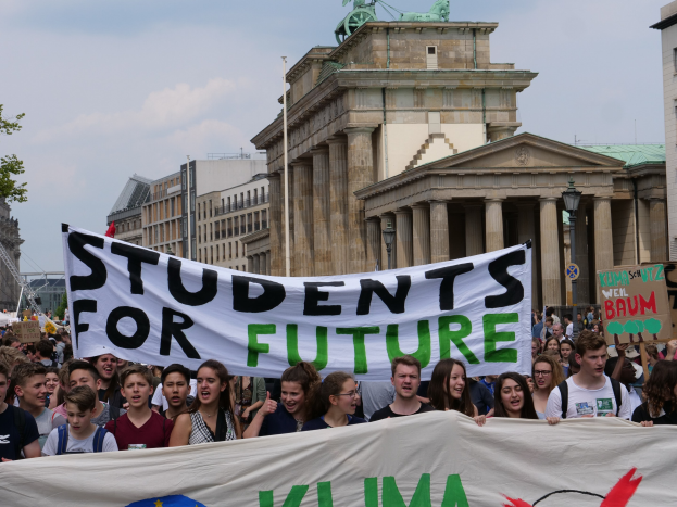 Gruppe von Schülern marschiert in Berlin mit einem bunt bemalten "Students for Future"-Schild vor einem Hintergrund aus Gebäuden, Bäumen und Himmel.