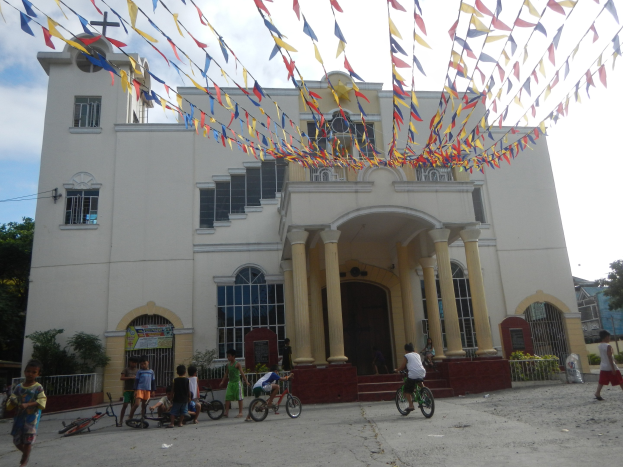 Eine Gruppe von Menschen, die vor einer weißen Kirche mit architektonischen Details wie Fenstern, Säulen, Bögen, Türen und einem Kreuzsymbol Fahrrad fahren, mit Pflanzen, einem Zaun, einer Fahne, Bäumen und einem bewölkten Himmel im Hintergrund und bunten Fahnen, die an einem Seil über ihnen befestigt sind.
