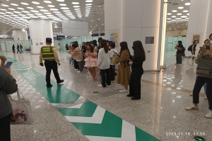 Eine Gruppe von Menschen steht in einer Reihe an einem Flughafen-Check-in, einige halten Taschen und Mobiltelefone, mit Säulen im Hintergrund und Lampen an der Decke und einem Wasserzeichen am unteren Bildrand.