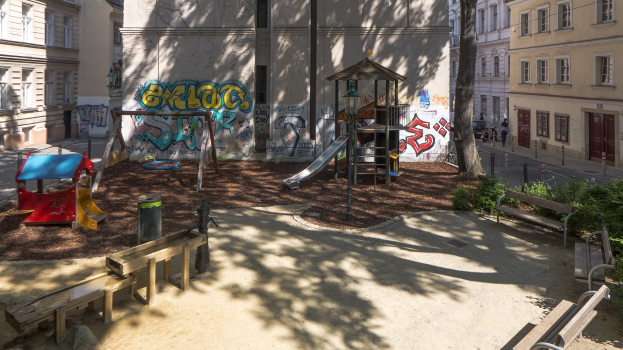 Spielplatz mit Graffiti, umgeben von städtischen Gebäuden, Bäumen, Bänken und einem Mülleimer, mit Menschen und Fahrzeugen auf der Straße und sichtbarem Himmel im Hintergrund.