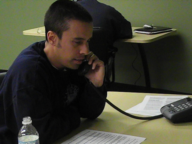 Mehrere Personen sitzen an einem Tisch mit einem Telefon, einer Einweg-Wasserflasche und Kabeln. Eine Person spricht am Telefon.